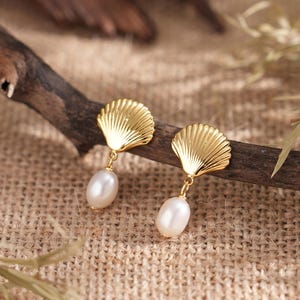 May include: A pair of gold-coloured seashell earrings with dangling white pearl drops. The earrings are displayed on a brown wooden branch against a neutral-toned textured background. The earrings have a classic, elegant design.