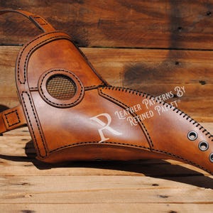 May include: A brown leather plague doctor mask with a long beak and a mesh eye covering. The mask has a strap that goes around the head and is decorated with stitching and eyelets. The text "Leather Patterns By Refined Parfait" is printed on the mask.