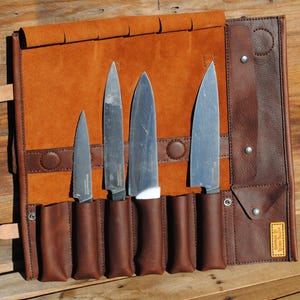 May include: A brown leather knife roll with four stainless steel knives. The roll is open, displaying the knives in individual pockets. The leather has a rich, warm tone, and the knives have silver blades and dark handles. The roll is secured with a leather strap.