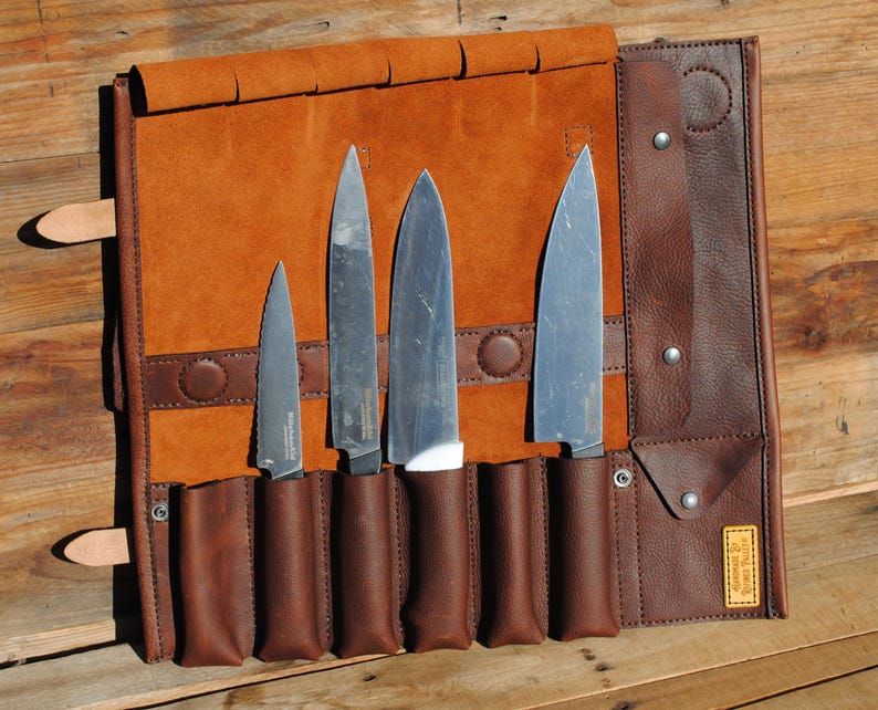 May include: A brown leather knife roll with four stainless steel knives. The roll is open, displaying the knives in individual pockets. The leather has a rich, warm tone, and the knives have silver blades and dark handles. The roll is secured with a leather strap.