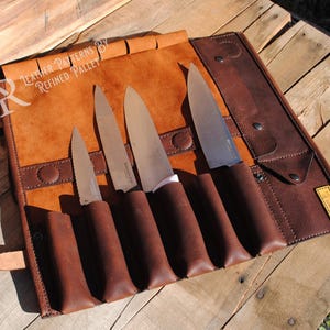 May include: A brown leather knife roll with four knives. The roll is made of a lighter brown leather with a darker brown leather trim and knife pockets. The knives have brown handles and silver blades. The text "LEATHER PATTERNS BY REFINED PALLET" is visible.