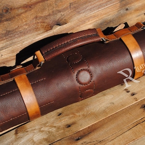 May include: A rolled-up, dark brown leather knife roll with tan leather straps and a handle. The roll is secured with two straps and features a stitched design. The text "Leather Patterns by Refined Pallet" is visible.