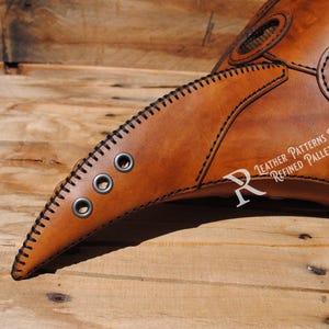 May include: A brown leather plague doctor mask with a long, curved beak and three metal eyelets. The mask has black stitching around the edges and is made by Refined Pallet.