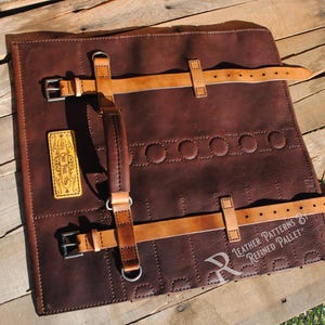 May include: A brown leather tool roll with tan leather straps and buckles. The roll has a stitched design and a small, rectangular patch with text. The roll is on a wooden surface.