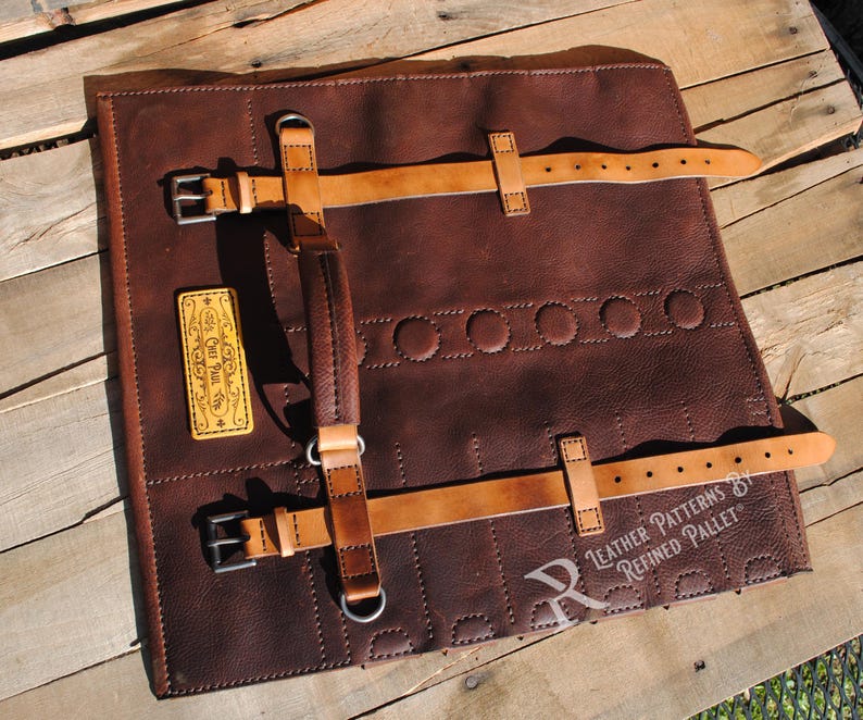 May include: A brown leather tool roll with tan leather straps and buckles. The roll has a stitched design and a small, rectangular patch with text. The roll is on a wooden surface.