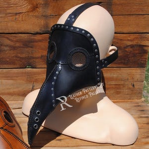 May include: A black leather plague doctor mask with a long beak and a mesh eye covering. The mask is worn on a mannequin head and has metal rivets and straps.