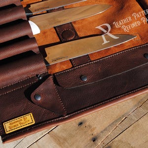 May include: A brown leather knife roll with several knives inside. The roll is made of dark brown leather and has a flap that secures with snaps. The knives have silver blades and white handles. The roll is on a wooden surface.
