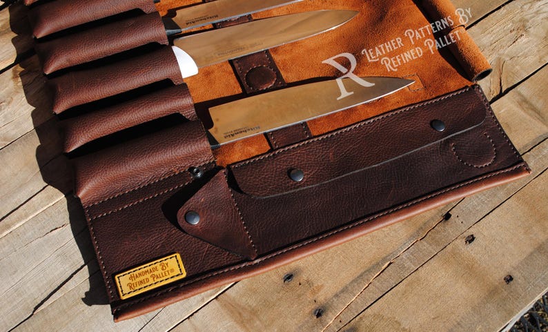 May include: A brown leather knife roll with several knives inside. The roll is made of dark brown leather and has a flap that secures with snaps. The knives have silver blades and white handles. The roll is on a wooden surface.