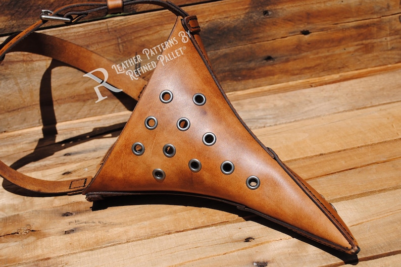 May include: A brown leather plague doctor mask with a long, pointed beak and multiple eyelets. The mask is attached to a brown leather strap. "Leather Patterns by Refined Pallet" is written on the strap.