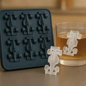 May include: A dark teal silicone ice cube tray with eight molds shaped like race cars. Two ice car cubes are outside the tray, and two are in a glass of amber liquid. The ice cubes are clear.