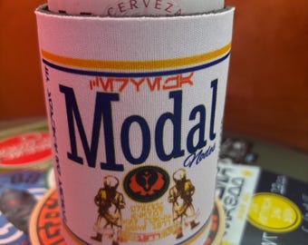 May include: A white beer can cooler with the word "Modal" in large blue letters. The cooler has a yellow and orange stripe design and features two figures in armor. The can contains "IMPORTED BEER" and "CERVEZA".