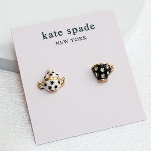 May include: A pair of gold-tone earrings featuring a black and white teapot and a black and white teacup with rhinestones. The earrings are on a pink card with the text "kate spade NEW YORK".
