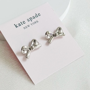 以下が含まれることがあります： シルバーカラーのリボン型スタッドピアス。ピアスは、黒で「kate spade NEW YORK」の文字が入ったピンク色のカードに表示されています。ピアスは光沢のある表面で、カードに配置されています。