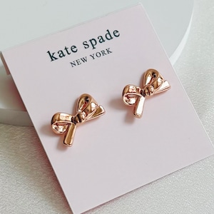 Puede incluir: Un par de pendientes en forma de lazo de oro rosa con el texto "kate spade new york" en una tarjeta rosa.