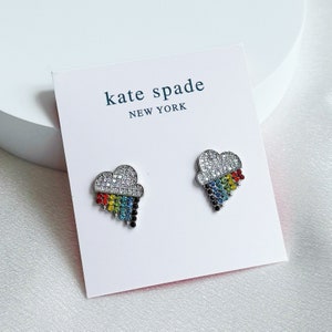 May include: A pair of Kate Spade cloud-shaped earrings. The top of the cloud is encrusted with clear crystals, and the bottom features a rainbow of colored crystals. The earrings are displayed on a white card with the Kate Spade New York logo.