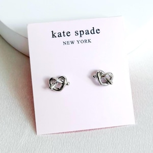 May include: Silver-toned pretzel stud earrings on a pink card with the "kate spade NEW YORK" logo. The earrings are small, with a knotted pretzel design. The background is white.