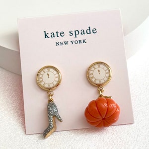 Op de afbeelding: Kate Spade oorbellen met een goudkleurige wijzerplaat met Romeinse cijfers. Eén oorbel heeft een zilveren glitter high heel bedel, de andere een gesneden oranje pompoen. De merknaam en locatie staan op de kaart.