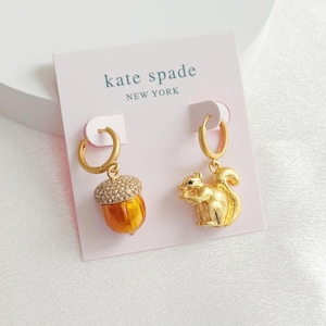 May include: Gold-tone hoop earrings. One earring features a detailed, gold-colored squirrel. The other earring has a gold-colored acorn with a crystal-studded cap. The earrings are displayed on a pink card with the text "kate spade NEW YORK".