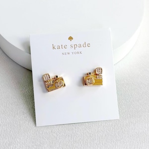 May include: Gold-toned camera stud earrings with clear crystal accents. The earrings are on a white card with the "kate spade NEW YORK" logo. The earrings are a fun accessory.