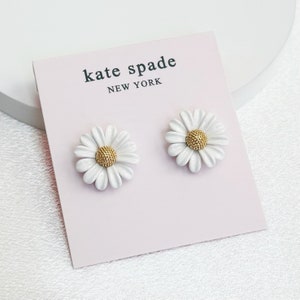 May include: A pair of white daisy stud earrings with gold-colored centers. The earrings are displayed on a pink card with the text "kate spade NEW YORK" in black.