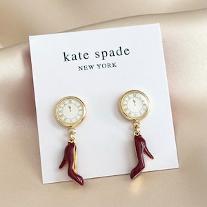 May include: A pair of gold-toned earrings featuring a clock face design with Roman numerals and a burgundy high-heeled shoe charm. The earrings are displayed on a white card with the text "kate spade NEW YORK".