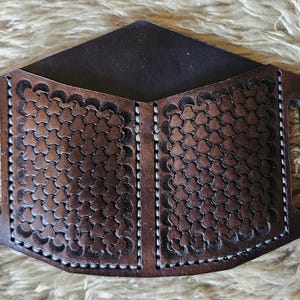 Puede incluir: Una cartera de cuero marrón con un patrón geométrico y un interior oscuro. La cartera tiene una forma única con asas recortadas a cada lado. El cuero está grabado con un diseño floral repetido y cosido con hilo blanco.