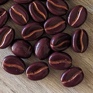 May include: A collection of dark burgundy coffee bean-shaped beads with a metallic gold-colored stripe. The beads are scattered on a light brown wooden surface. The beads have a glossy finish.