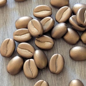 May include: A collection of metallic gold-colored coffee bean beads. Some beads are strung on a silver-colored wire, while others are scattered on a light wood-grain surface. The beads have a detailed, realistic coffee bean shape, perfect for jewelry making or craft projects.
