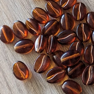 May include: A collection of brown, coffee bean-shaped beads with a glossy finish. Each bead has a lighter, gold-colored stripe down the center, creating a realistic coffee bean appearance. The beads are scattered on a light brown surface.
