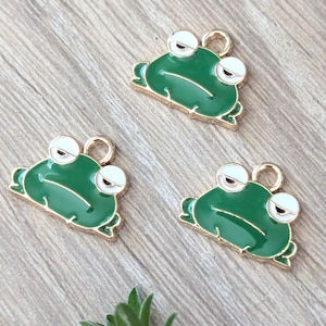 Puede incluir: Tres dijes de rana verdes con contornos dorados y ojos blancos. Cada dije tiene un lazo dorado en la parte superior para sujetarlo a joyas u otras manualidades. Los dijes están sobre una superficie de madera marrón claro.