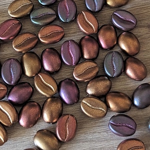 May include: A collection of iridescent coffee bean shaped beads in shades of brown, purple, and green. The beads are arranged in a random pattern on a wooden surface.