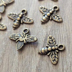 May include: Close-up of several small, bronze-colored bee charms. Each charm features detailed wings, a segmented body, and a small loop for attaching to jewelry or other crafts. The charms are scattered on a light brown surface.