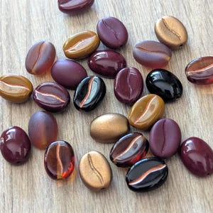 May include: A collection of coffee bean-shaped beads in various colors, including shades of brown, gold, black, and burgundy. The beads have a glossy or translucent finish, with some featuring stripes.