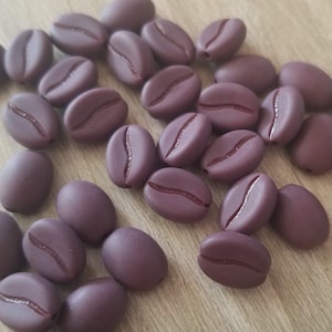 Puede incluir: Una colección de cuentas de color marrón oscuro con forma de grano de café. Las cuentas están hechas de un material liso y mate y tienen un diseño ligeramente elevado en la superficie.