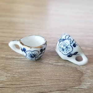 May include: Two miniature white teacups with blue and gold floral designs. The cups are on a wooden surface.