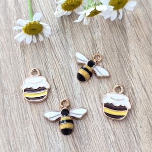 May include: Four gold-toned bee and cupcake charms with white, black, and yellow enamel details. The charms are scattered on a wooden surface with daisy flowers.