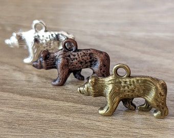 May include: Three small bear charms in silver, bronze, and brass tones are arranged diagonally on a wooden surface. Each bear has a loop at the top for hanging. The charms are detailed with fur and bear features.