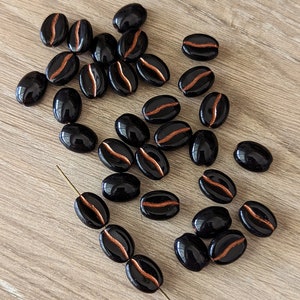 May include: A collection of black and copper coffee bean shaped beads. The beads are oval shaped and have a copper stripe down the center.