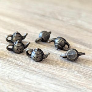 May include: A collection of small, antique-style bronze teapots. Each teapot features a handle, spout, and lid, with decorative cutouts on the body. The teapots are arranged on a light-colored wooden surface.