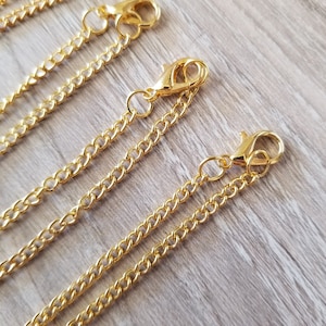 May include: Close-up of several gold-colored chain necklaces with lobster claw clasps. The chains are made of small, interlocking links. The clasps are attached to the chains with small jump rings. The background is a light wood grain.