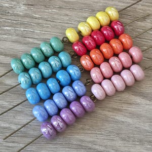 May include: A collection of smooth, round, glossy ceramic beads arranged in rows on thin metal wires. The beads feature a vibrant gradient of colors including yellow, red, orange, pink, light blue, teal, and purple, each with a reflective finish.