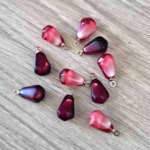 May include: A collection of nine pomegranate-shaped pendants. The pendants are a mix of deep red and pink, with a gold-colored loop at the top for attaching to a chain or other jewelry component. The pendants are scattered on a light wood surface.