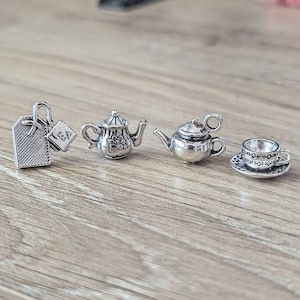 May include: Four silver charm pendants: a tea bag tag with the word "TEA", a detailed teapot with a floral design, a simple teapot, and a teacup and saucer.