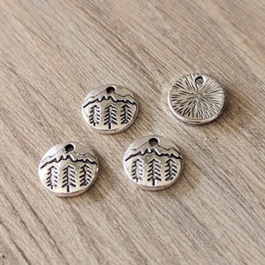 May include: Four silver charm pendants, three feature a mountain range with a sun and pine trees, one has a sunburst design.