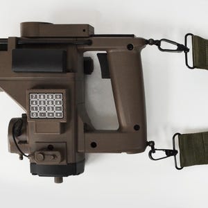 May include: A brown and black device with a keypad and a black handle. It has a green strap attached with black clips. The device appears to be a futuristic or tactical tool.