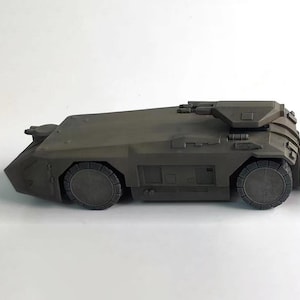 Puede incluir: Modelo gris oscuro de un vehículo blindado de perfil bajo. El vehículo tiene un frente inclinado, una torreta con un cañón y ruedas circulares. El modelo está detallado con líneas de panel y otras características.