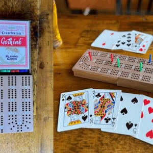 Op de afbeelding: Een verzameling spellen op een houten tafel. Een pakje "Gothink" speelkaarten, een cribbagebord met pinnen en speelkaarten zijn zichtbaar. De speelkaarten tonen verschillende kleuren en getallen. Het cribbagebord heeft rode, groene en blauwe pinnen.