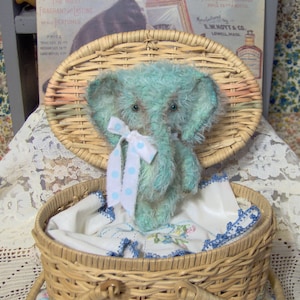 Puede incluir: Un juguete de peluche de elefante azul claro con un lazo blanco y lunares azules se sienta dentro de una cesta de mimbre con un forro de tela blanca. La cesta tiene una tapa y un asa.
