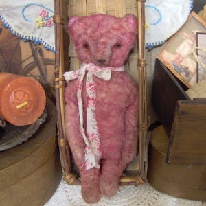May include: A vintage-style pink teddy bear with a floral ribbon around its neck. The bear is seated in a small, woven chair. The bear has a distressed appearance, suggesting it is an antique or handmade item.
