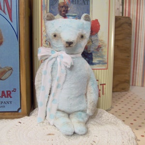May include: A light blue teddy bear with a white and blue polka dot scarf. The bear has brown eyes and a stitched nose. It is sitting on a white lace doily. The background includes vintage toy packaging.
